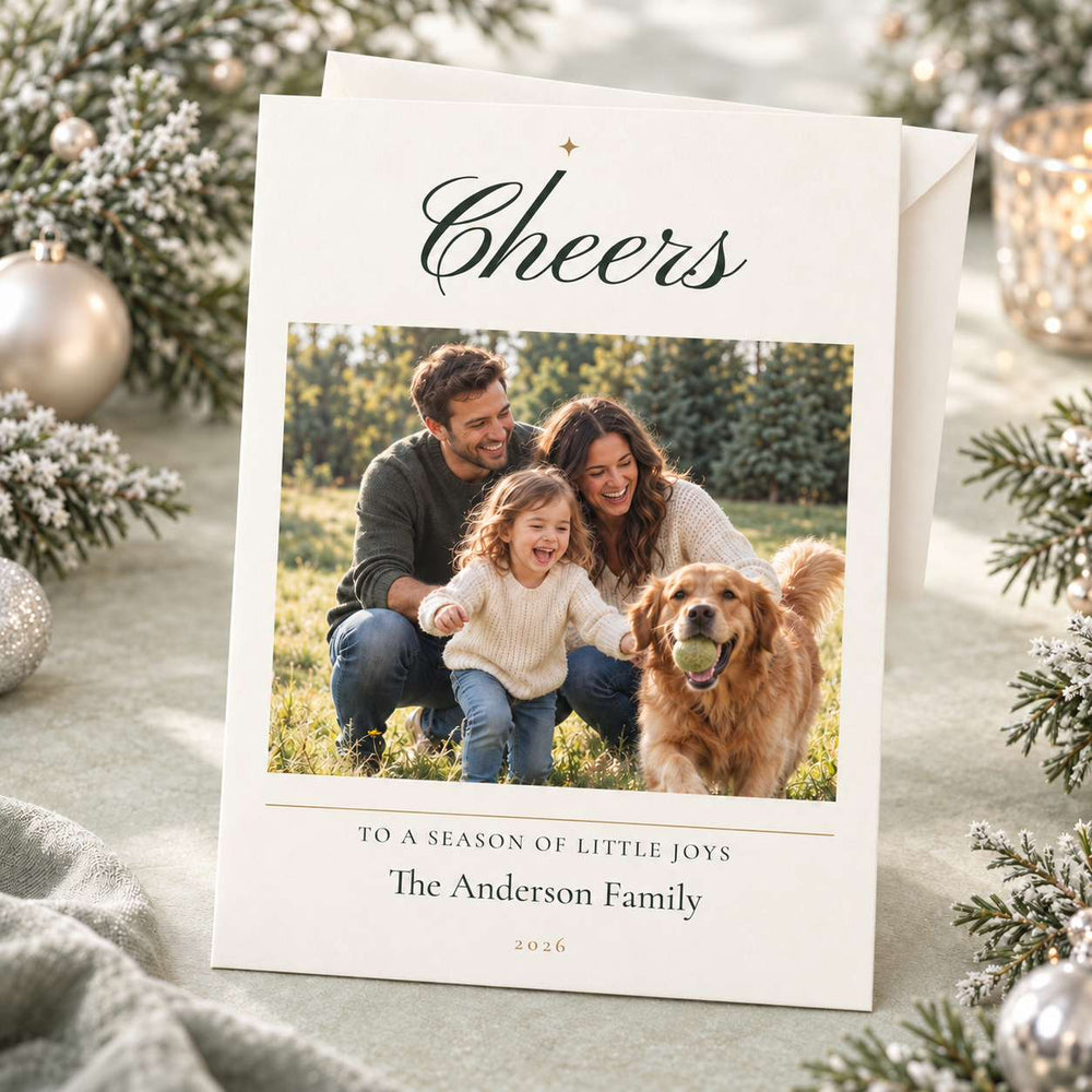 A personalized Christmas photo card with an envelope, styled on a soft gray fabric with holiday greenery and ornaments. The card features the word 'Cheers' at the top and a photo of a family and their golden retriever dog in the center, with the text 'TO A SEASON OF LITTLE JOYS The Anderson Family 2026' printed below.