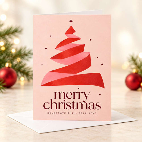 A pink Christmas greeting card with a stylized red paper-cut tree and the words “merry christmas” and “celbrate the little joys,” shown standing on a white envelope with blurred holiday greenery and red ornaments in the background.