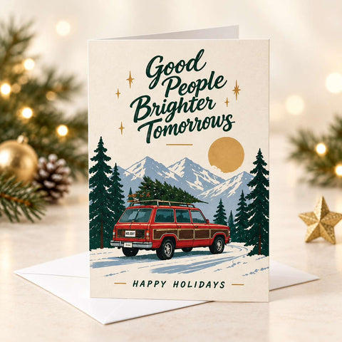 A holiday greeting card with a white envelope, styled on a wooden table with Christmas decorations including pine branches, a lantern, wrapped gifts, and a mug of hot chocolate. The card features a cream background with an illustration of a red station wagon driving through a snowy mountain landscape under a full moon, with the text 'Good People Brighter Tomorrows' and 'HAPPY HOLIDAYS'.